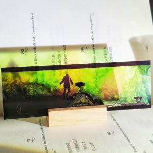 K&ouml;nnte beinhalten: Ein rechteckiger Lesezeichen mit einer Silhouette einer Person in der N&auml;he einer Struktur, vor einem Hintergrund aus gr&uuml;nen und gelben Farbt&ouml;nen. Das Lesezeichen wird von einem kleinen, rechteckigen Holzblock getragen, der auf den Seiten eines Buches liegt.