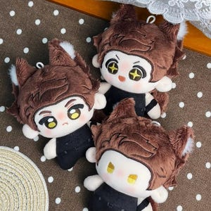 May include: Three plush dolls featuring brown fur, pointed animal ears with white tips, and black outfits. Each doll has embroidered facial features, including large eyes with yellow accents, pink cheeks, and a small mouth, with a small loop on top.