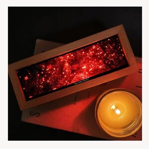 K&ouml;nnte beinhalten: Ein rechteckiges Kunstwerk mit Holzrahmen, das eine rote Galaxieszene mit dem Text "HAIL MARY PROJECT" zeigt. Eine brennende Kerze in einem Glasgef&auml;&szlig; befindet sich in der N&auml;he und strahlt ein warmes Licht aus. Das Kunstwerk liegt auf einer roten Oberfl&auml;che.