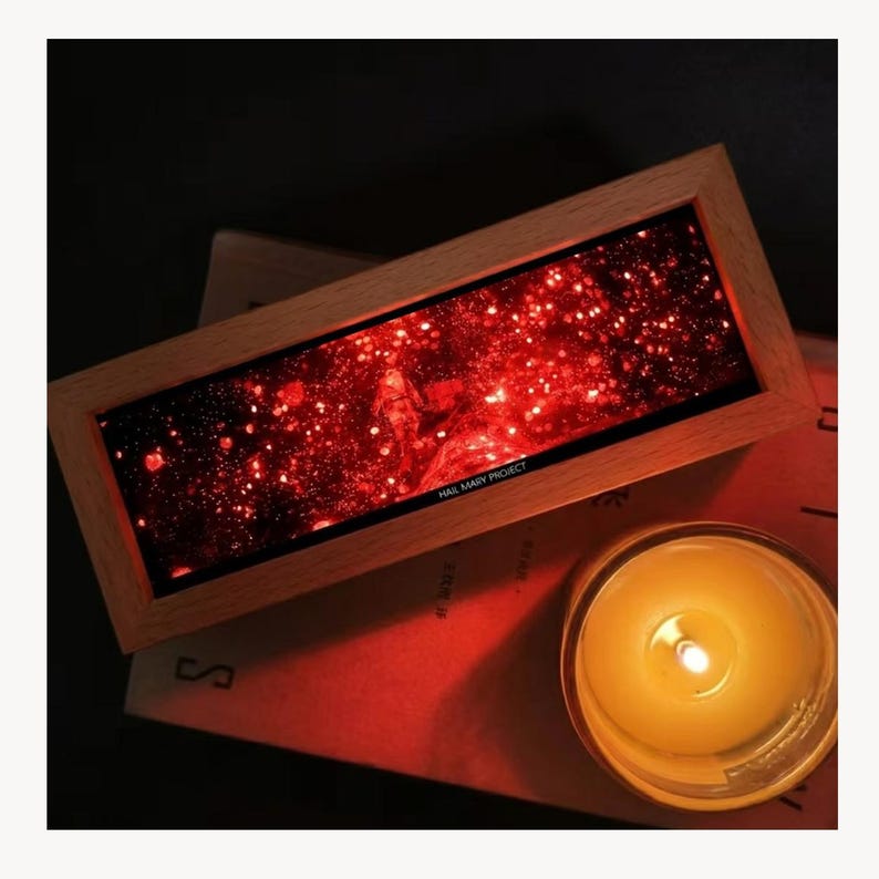 K&ouml;nnte beinhalten: Ein rechteckiges Kunstwerk mit Holzrahmen, das eine rote Galaxieszene mit dem Text "HAIL MARY PROJECT" zeigt. Eine brennende Kerze in einem Glasgef&auml;&szlig; befindet sich in der N&auml;he und strahlt ein warmes Licht aus. Das Kunstwerk liegt auf einer roten Oberfl&auml;che.