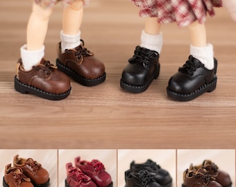 May include: Miniature doll shoes in brown and black leather. The shoes have laces and thick soles. The image also shows a pair of red shoes. The shoes are displayed on a wooden surface.