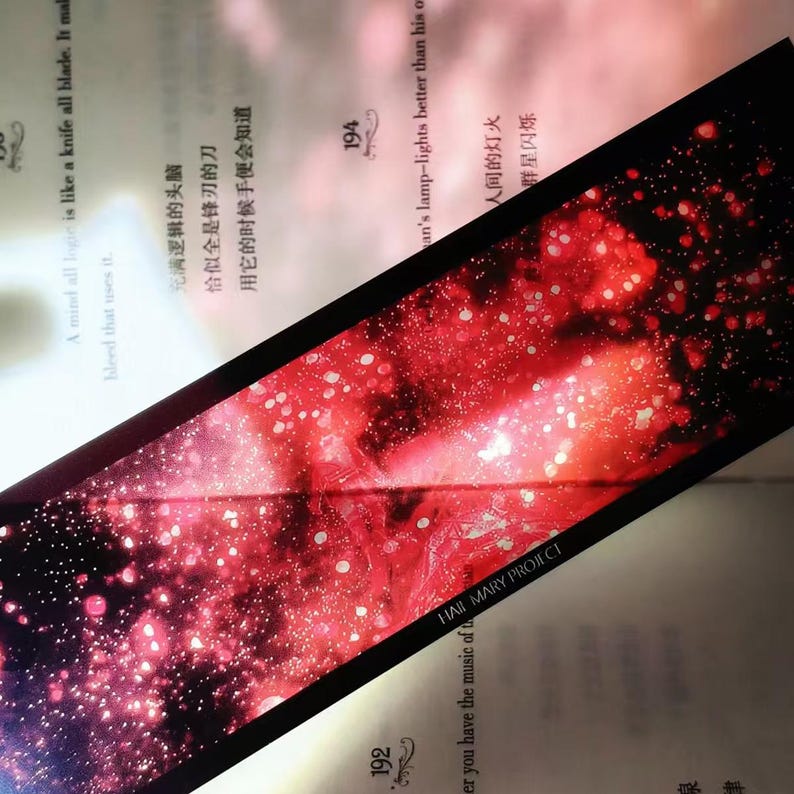 K&ouml;nnte beinhalten: Ein rechteckiger Lesezeichen mit einem roten und wei&szlig;en Galaxiemuster. Der Lesezeichen hat einen schwarzen Rand und der Text "HAJI MARY PROJECT" ist sichtbar. Es liegt auf einem offenen Buch.