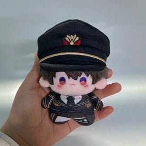 Op de afbeelding: Een pluche speelgoed van een personage dat een zwarte pet draagt met een gouden rand en een rood en gouden embleem. Het personage draagt een zwart en gouden uniform met een witte kraag en een zwarte stropdas. Het speelgoed heeft bruin haar en blauwe ogen.
