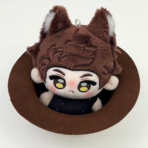 May include: A plush doll with brown hair and wolf-like ears featuring white tips, large yellow eyes, and a small mouth, wearing a black outfit. The doll is positioned inside a brown circular hat, which has a small silver loop at the top.