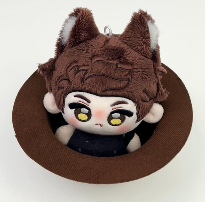 May include: A plush doll with brown hair and wolf-like ears featuring white tips, large yellow eyes, and a small mouth, wearing a black outfit. The doll is positioned inside a brown circular hat, which has a small silver loop at the top.