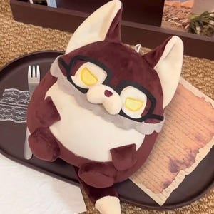 May include: A plush toy shaped like a round fox with dark maroon fur, a cream-colored belly, and a white-tipped tail. The fox features large, pointed ears, a small nose, and black-rimmed glasses over yellow eyes with a grey ruff around the neck.