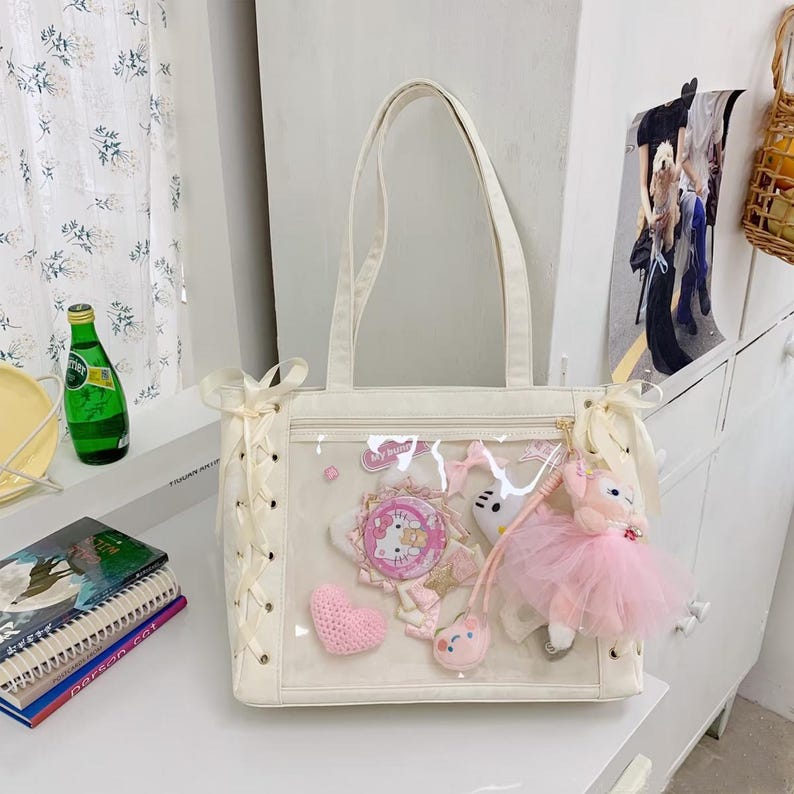 Pode incluir: Bolsa tote creme com um painel frontal transparente exibindo itens decorativos. Inclui um cora&ccedil;&atilde;o rosa, um distintivo Hello Kitty e uma boneca de pel&uacute;cia rosa com tutu. A bolsa tem al&ccedil;as creme e detalhes de cadar&ccedil;o.