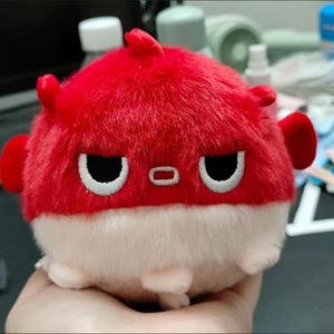 May include: A red and white plush toy shaped like a pufferfish with a grumpy expression. The toy has black eyes and a small red mouth.
