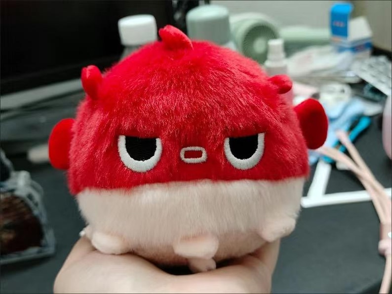 May include: A red and white plush toy shaped like a pufferfish with a grumpy expression. The toy has black eyes and a small red mouth.