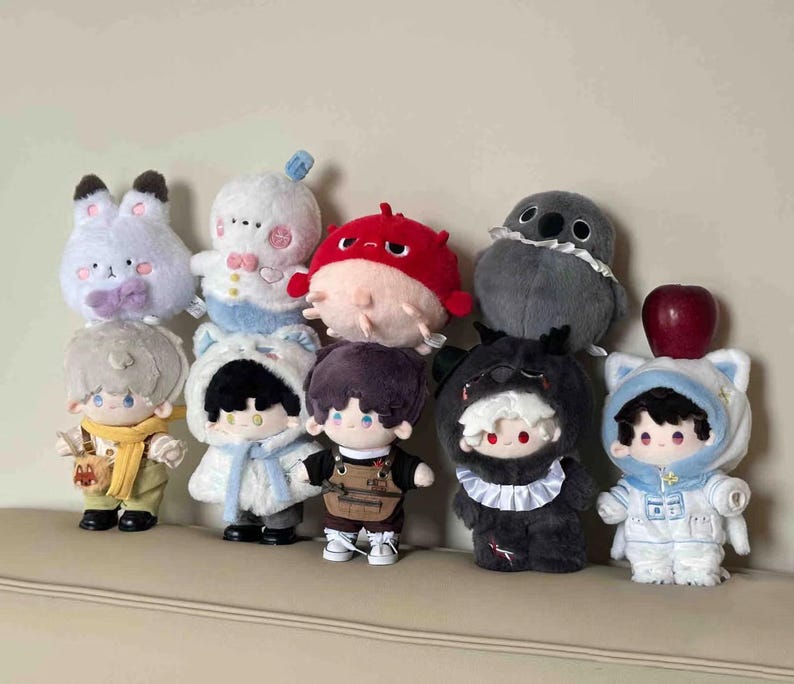 K&ouml;nnte beinhalten: Eine Sammlung von Pl&uuml;schpuppen in verschiedenen Tier- und Charakterkost&uuml;men. Die Puppen haben detaillierte Gesichtsausdr&uuml;cke und tragen Outfits wie ein Hase, ein Kugelfisch und ein B&auml;r. Ein roter Apfel liegt auf einer der Puppen.