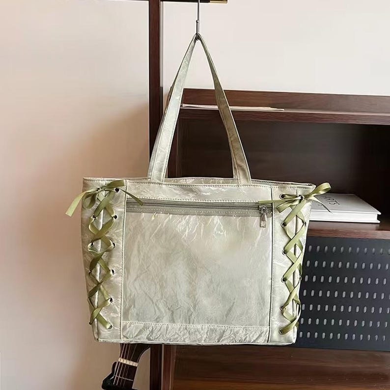 Pode incluir: Uma sacola verde clara com fecho de z&iacute;per e detalhes em fita verde. A sacola tem duas al&ccedil;as e est&aacute; pendurada em um gancho de metal. A sacola &eacute; feita de um material transl&uacute;cido.