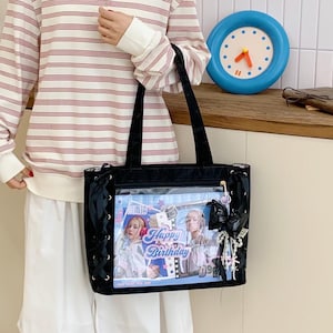 Pode incluir: Bolsa tote preta com um bolso frontal transparente exibindo um gr&aacute;fico "Happy Birthday" com duas mulheres. A bolsa tem detalhes em fita preta e um charme decorativo. A bolsa &eacute; segurada por uma pessoa vestindo uma camisa listrada.