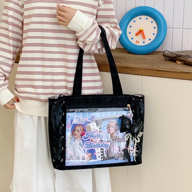Pode incluir: Bolsa tote preta com um bolso frontal transparente exibindo um gr&aacute;fico "Happy Birthday" com duas mulheres. A bolsa tem detalhes em fita preta e um charme decorativo. A bolsa &eacute; segurada por uma pessoa vestindo uma camisa listrada.