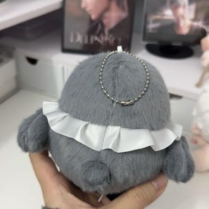 May include: A gray plush toy with a white ruffle around the bottom. The toy has a silver chain attached to it.