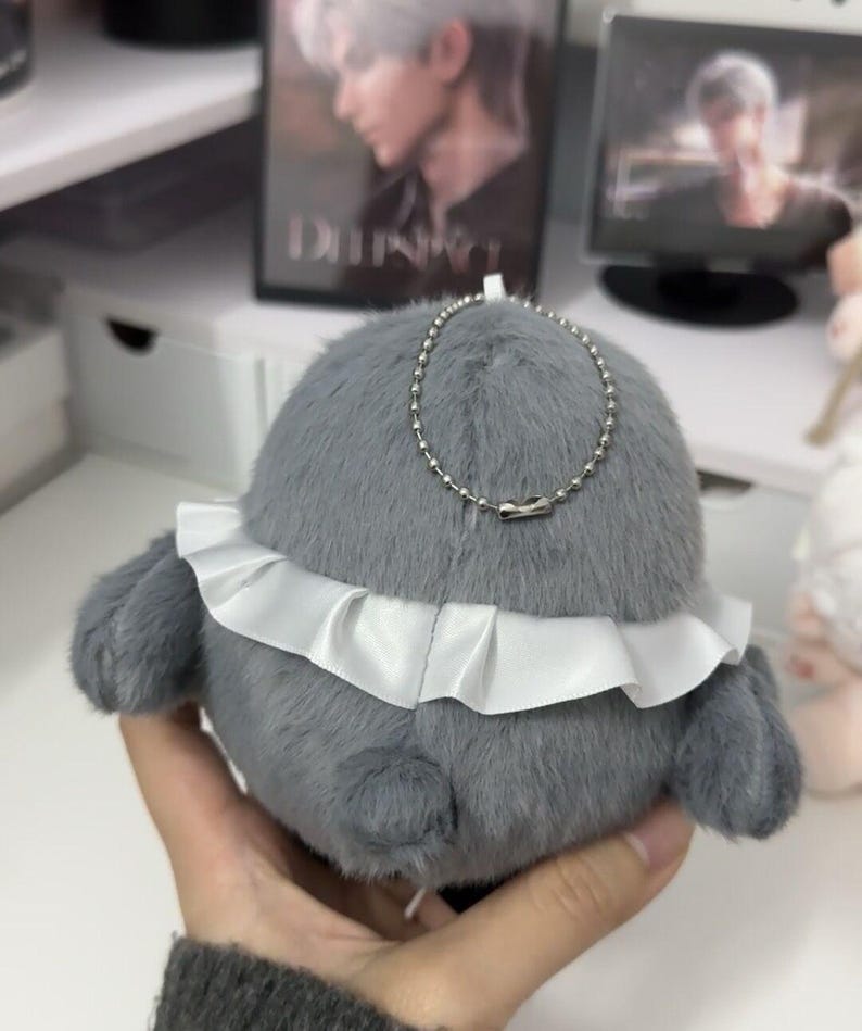 May include: A gray plush toy with a white ruffle around the bottom. The toy has a silver chain attached to it.