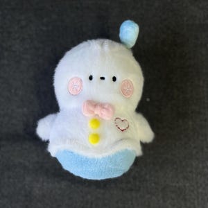 May include: A white plush toy with a blue hat, pink cheeks, and a yellow bow tie. The toy has a heart-shaped patch on its chest and two yellow buttons.