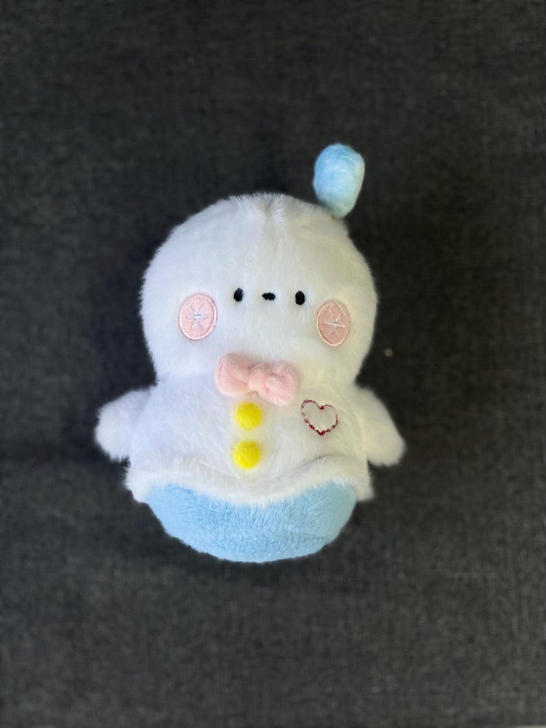 May include: A white plush toy with a blue hat, pink cheeks, and a yellow bow tie. The toy has a heart-shaped patch on its chest and two yellow buttons.