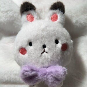 May include: A white plush bunny with black ears and pink cheeks. The bunny is wearing a purple bow tie.