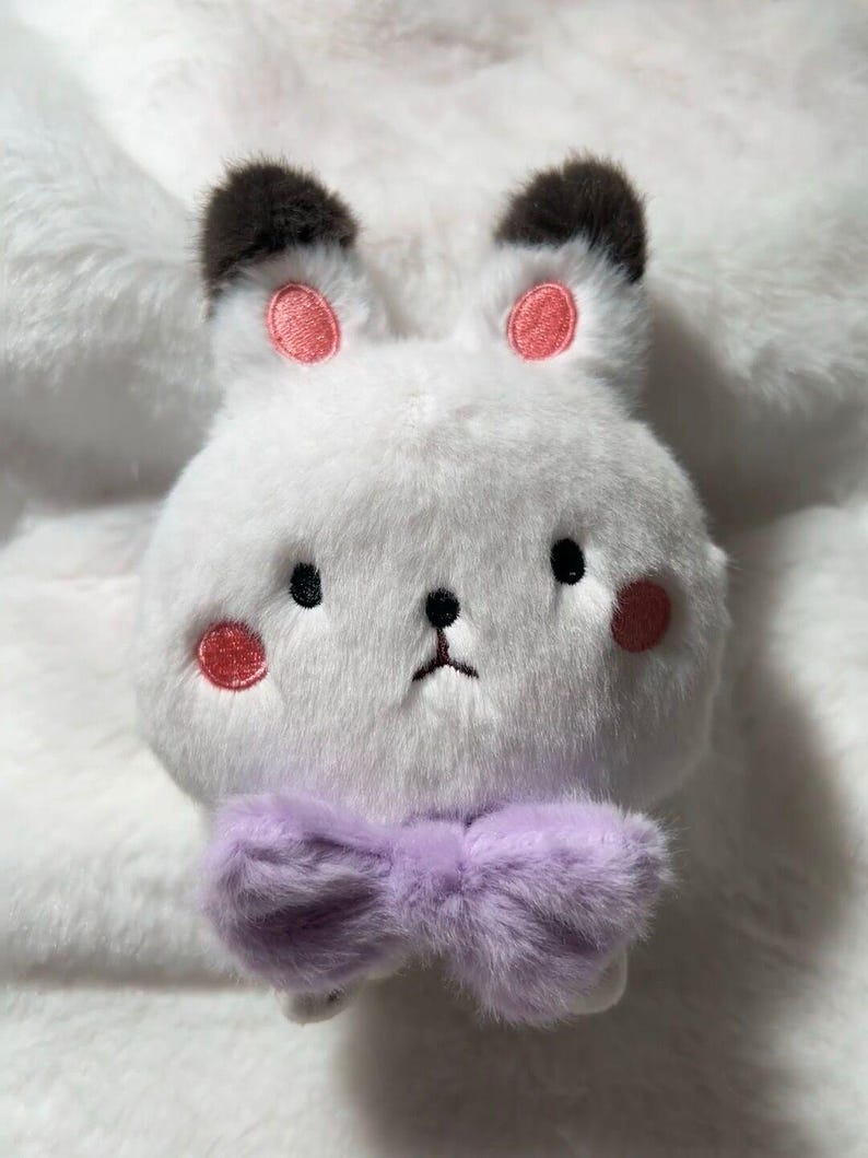 May include: A white plush bunny with black ears and pink cheeks. The bunny is wearing a purple bow tie.