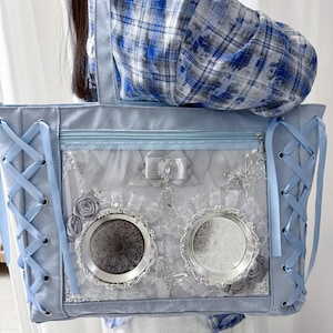 Pode incluir: Uma mala tote azul clara com um design decorativo. A mala apresenta um detalhe de atacadores nas laterais, um fecho de correr na parte superior e um painel transparente com enfeites prateados, incluindo duas molduras redondas e ornamentadas. Rosas cinzentas e um la&ccedil;o completam o design.