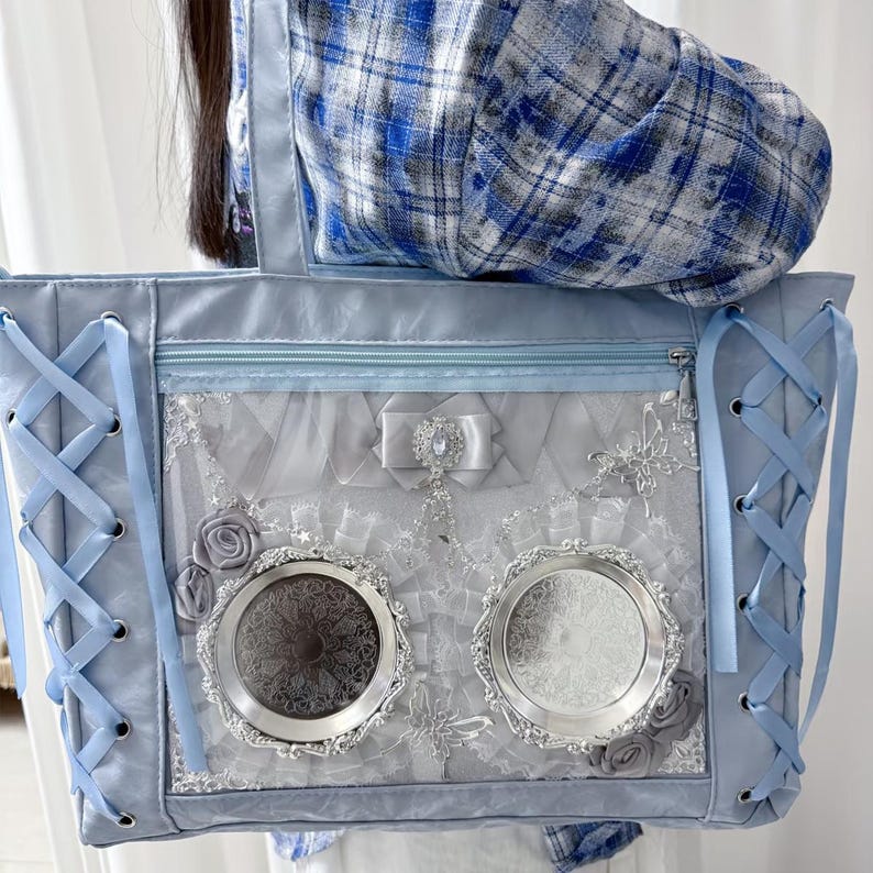 Pode incluir: Uma mala tote azul clara com um design decorativo. A mala apresenta um detalhe de atacadores nas laterais, um fecho de correr na parte superior e um painel transparente com enfeites prateados, incluindo duas molduras redondas e ornamentadas. Rosas cinzentas e um la&ccedil;o completam o design.