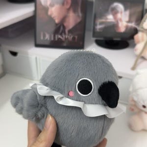 May include: A gray plush bird with a white ruffle collar and black eyes. The bird has a small pink blush on its cheek and a black beak.