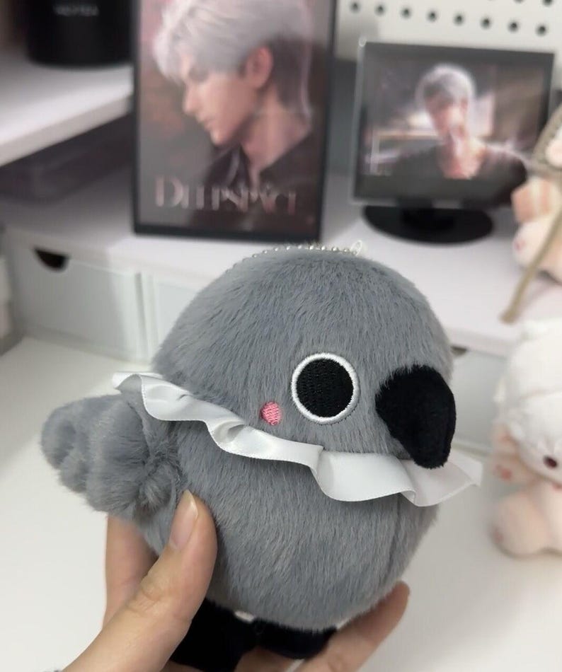May include: A gray plush bird with a white ruffle collar and black eyes. The bird has a small pink blush on its cheek and a black beak.