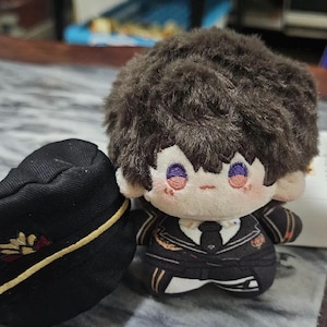 Op de afbeelding: Een pluche speelgoed van een personage met bruin haar, dat een zwart en gouden uniform met een stropdas draagt. Het speelgoed heeft een kleine zwarte hoed met gouden versieringen.