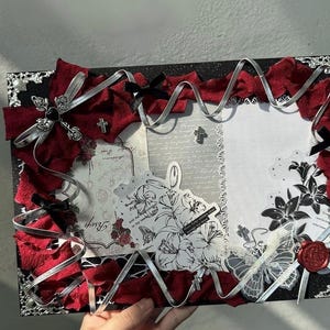 Pode incluir: Uma moldura decorativa com uma borda preta brilhante, adornada com tecido vermelho, fita prateada e la&ccedil;os pretos. A moldura apresenta ilustra&ccedil;&otilde;es florais e de borboletas, cruzes e um selo de cera vermelho.