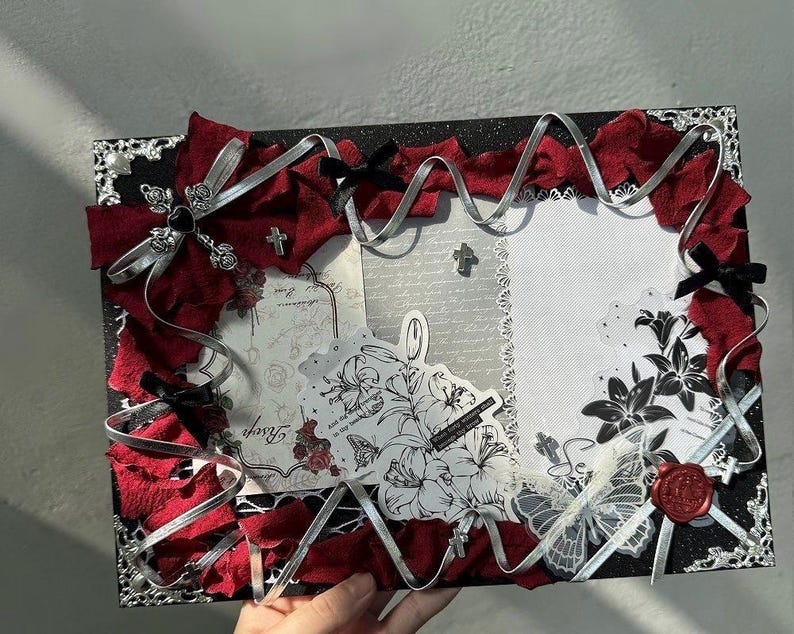 Pode incluir: Uma moldura decorativa com uma borda preta brilhante, adornada com tecido vermelho, fita prateada e la&ccedil;os pretos. A moldura apresenta ilustra&ccedil;&otilde;es florais e de borboletas, cruzes e um selo de cera vermelho.