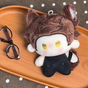 May include: A plush doll with brown fur, pointed ears, and yellow eyes, wearing a black outfit. The doll has a white face with pink cheeks and a small metal loop on top. Beside the doll are round, brown-rimmed glasses with dark temples.
