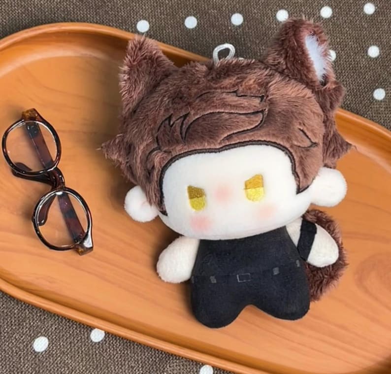 May include: A plush doll with brown fur, pointed ears, and yellow eyes, wearing a black outfit. The doll has a white face with pink cheeks and a small metal loop on top. Beside the doll are round, brown-rimmed glasses with dark temples.