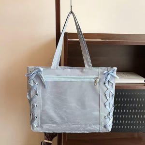 Pode incluir: Uma mala tote azul clara com detalhes de fita nas laterais. A mala tem fecho de correr e al&ccedil;as compridas. A mala est&aacute; pendurada num gancho de metal.