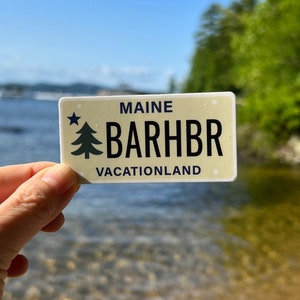 May include: A yellow and white sticker with a blue star and pine tree design. The sticker says "Maine" and "BARHBR" with the text "VACATIONLAND" below.