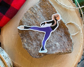 Könnte beinhalten: Ein Aufkleber mit einer Frau, die Schlittschuh läuft, in einem lila und schwarzen Outfit auf einem braunen Felsen. Der Aufkleber befindet sich auf einer Holzoberfläche mit einem Stück Schnur und einer Schnurrolle.