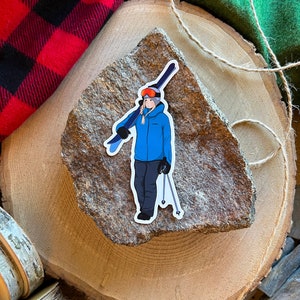 May include: A sticker of a person wearing a blue jacket and a ski helmet holding skis. The sticker is on a brown rock.