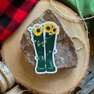 May include: A sticker of green rain boots with sunflowers and vines growing out of the tops of the boots. The sticker is on a brown rock.