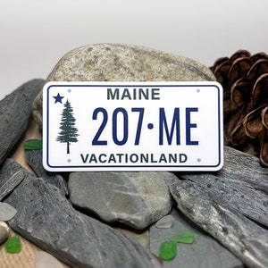 May include: A white license plate-shaped sticker with the text "MAINE 207•ME VACATIONLAND". The sticker features a green pine tree and a blue star. The background includes gray stones, green glass pieces, and a pine cone.