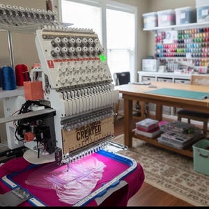 May include: A large, white embroidery machine with multiple spools of thread. The machine is actively embroidering a pink fabric, with the text "Family is Created" visible. The workspace includes spools of colourful thread and a wooden table.