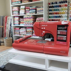 May include: A red sewing machine, model EOC05-RE, is on a white table in a sewing room. The machine has a display with the text "Contact Us to Be An Expert". Shelves behind the machine are filled with colourful fabric, and spools of thread are on the wall.