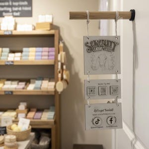 May include: A wooden sign with the text "SERENITY ACRES GOATS" and a goat illustration hangs from a wooden rod. Below, a "SCAN TO PAY" sign and a social media sign with the text "Let's get Social!" are visible.