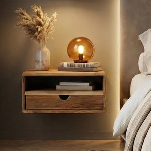 Puede incluir: Una mesita de noche flotante de madera con un cajón y un estante, con libros, un jarrón decorativo con flores secas y una lámpara esférica de cristal ámbar. Una lámpara colgante gris cuelga encima. La escena es en un dormitorio.