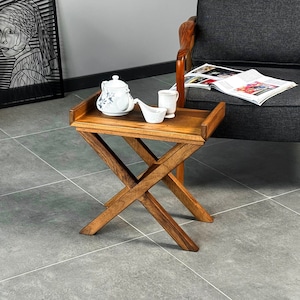 Può includere: Un tavolo pieghevole in legno con finitura marrone. Il tavolo ha una base a forma di croce e un piano rettangolare. Un bollitore bianco, una tazza bianca e un piattino bianco sono sul tavolo.