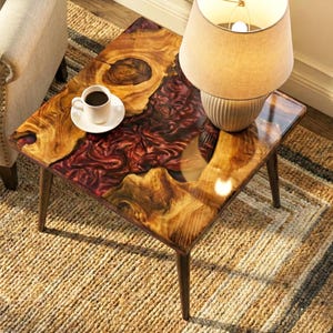 May include: A small, square coffee table with a unique wood and resin design. The tabletop features a mix of warm wood tones and deep red resin, with a clear glossy finish. A beige lamp with a round lampshade sits on the table, next to a white coffee cup.