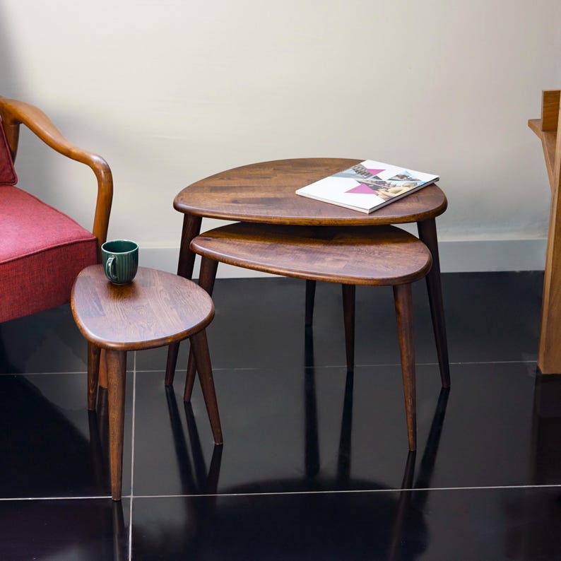 May include: Set of three mid-century modern nesting tables. The tables have a dark wood finish and a unique, organic shape. The largest table has a magazine on top. The tables are supported by slender, tapered legs. A green mug sits on the smallest table.