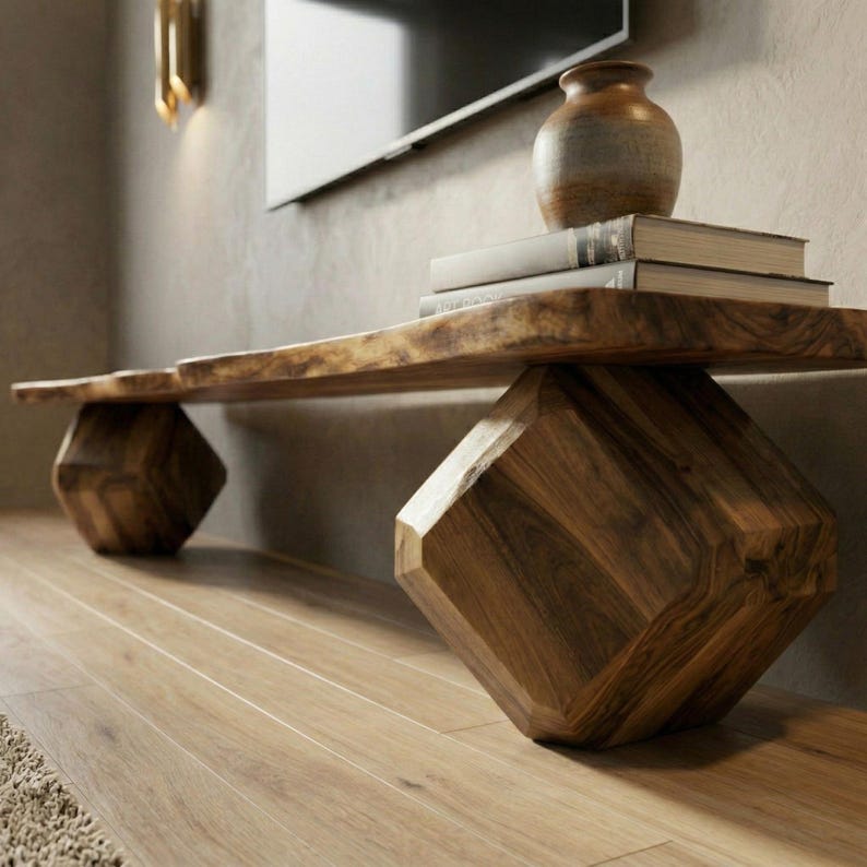 May include: A long wooden bench with a natural live edge top and two large, geometric, faceted cube-shaped legs. A small, rounded, earth-toned ceramic vase sits on top of two stacked books placed on the surface of the wooden bench.