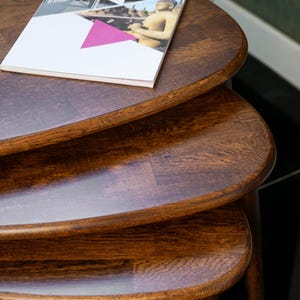 May include: A set of three nesting tables made of dark brown wood. The tables are oval-shaped with rounded edges and a glossy finish. A magazine with a white cover and a pink triangle is on the top table.