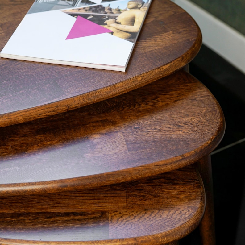 May include: A set of three nesting tables made of dark brown wood. The tables are oval-shaped with rounded edges and a glossy finish. A magazine with a white cover and a pink triangle is on the top table.