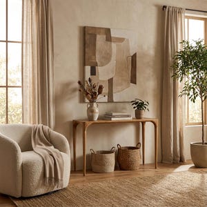 May include: A cozy living room scene with neutral tones. A curved wooden console table holds a vase of dried flowers, books, and a potted plant. Two woven baskets sit below. A cream-colored armchair with a blanket and a large abstract painting complete the decor.
