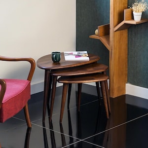 May include: Set of three nesting tables in a warm brown finish. The tables have a rounded, organic shape and tapered legs. A dark green mug and a magazine rest on the top table. The tables are a stylish addition to any living space.